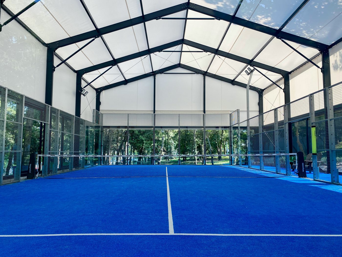 A bright indoor padel facility with a glass-roof structure and a blue court